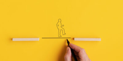 Wide view image of male hand drawing a line between two wooden pegs for a silhouette of a man to walk across. Over yellow background with copy space.